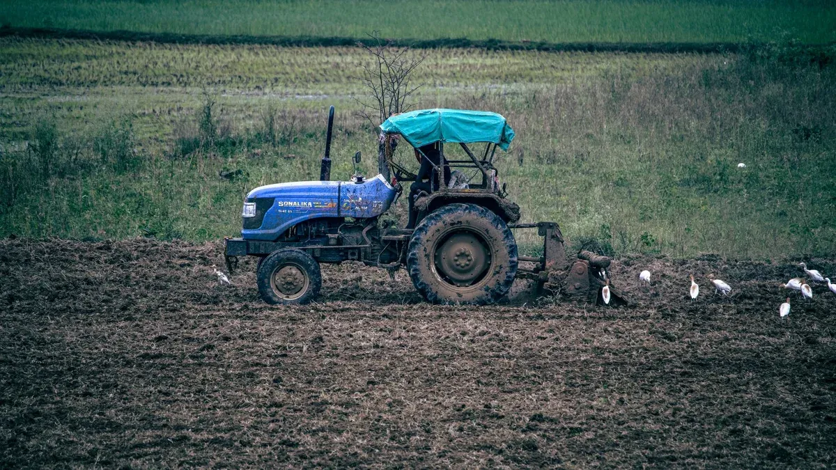 A blue tractor plowing a field in rural India with birds flocking around.