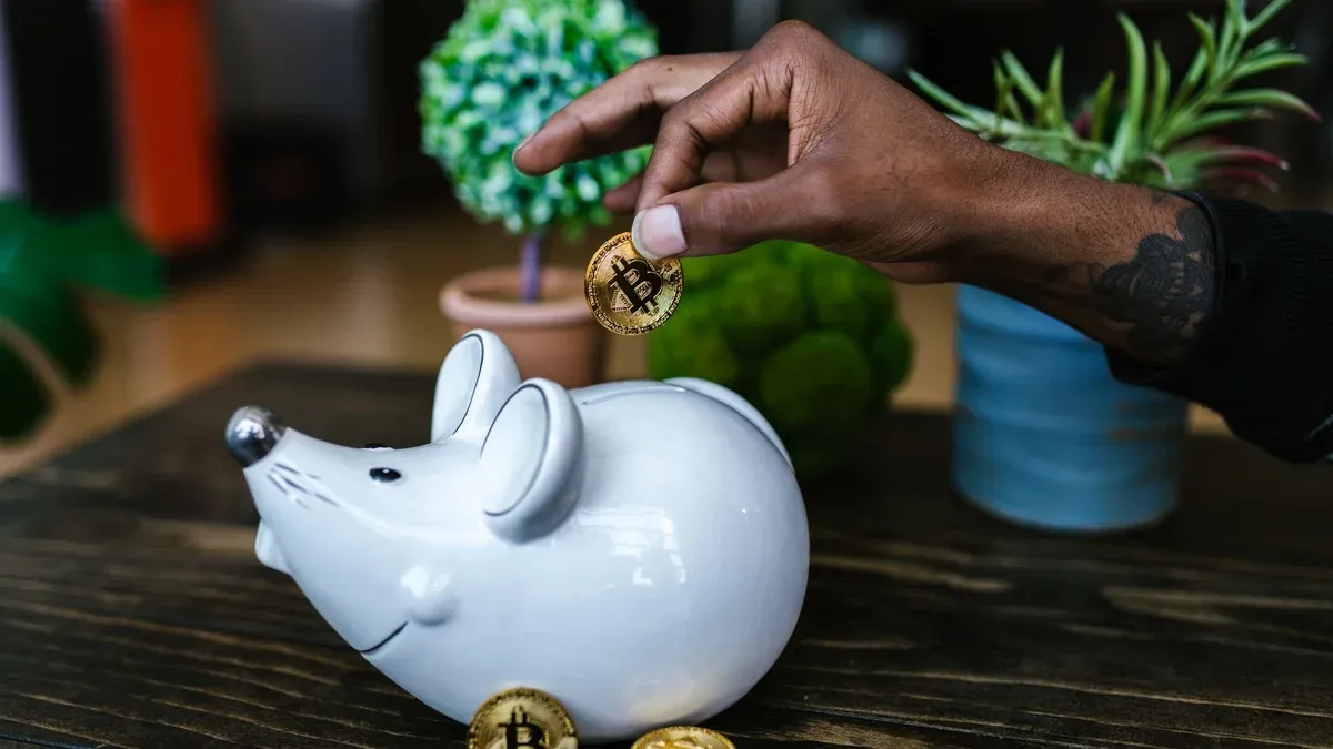 Close-up of hand placing bitcoin into a mouse-shaped piggy bank on a table.