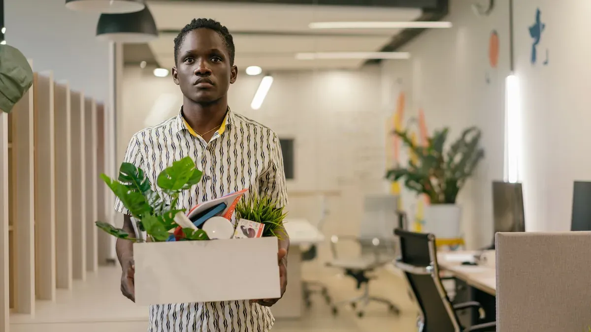 A serious man carries a box of belongings in an office setting, symbolizing dismissal or job change.