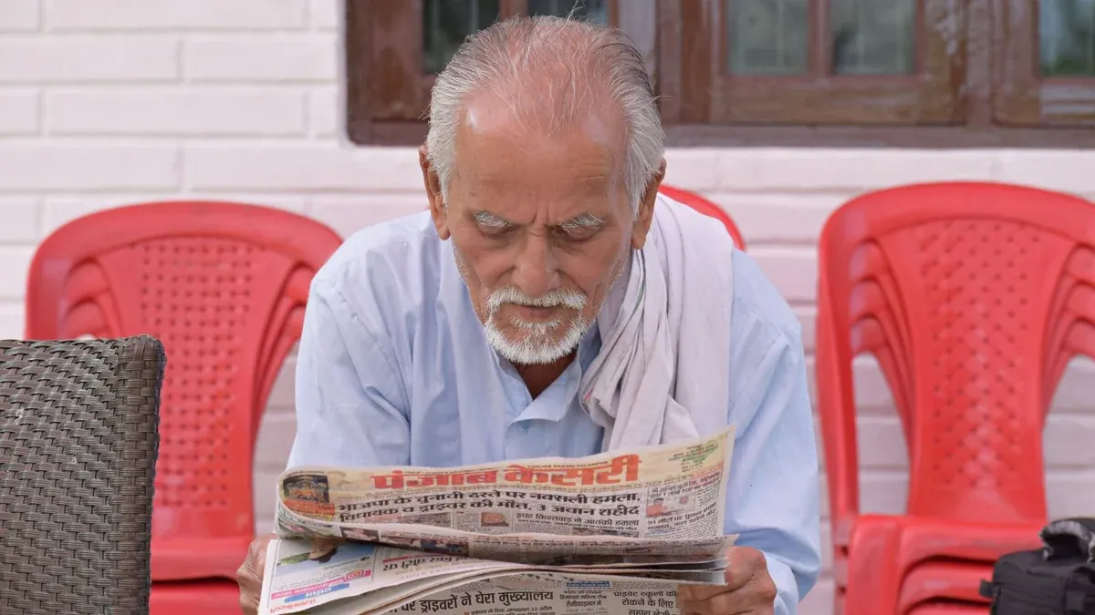 Elderly man engrossed in reading a newspaper while seated outside with red chairs around.