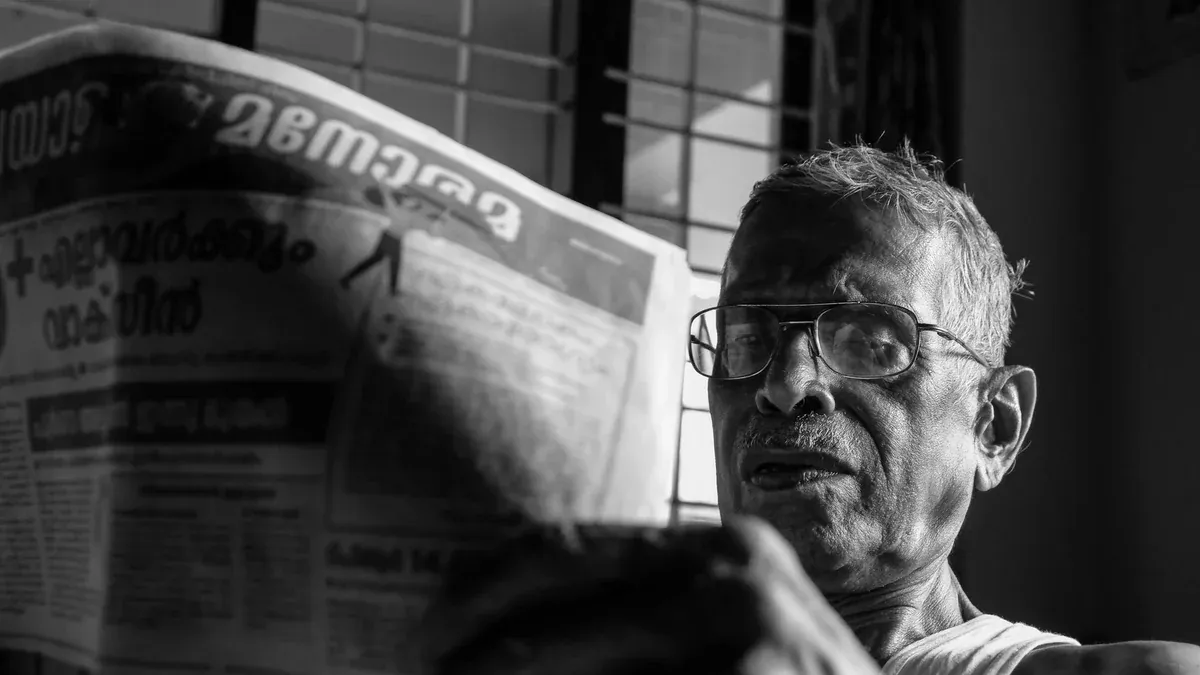 Black and white photo of an elderly man with glasses reading a newspaper in Nilambur, India.