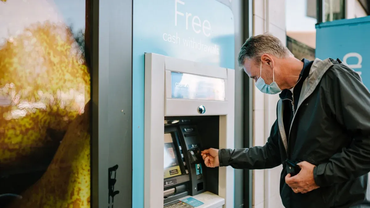 A man wearing a mask using an ATM machine outdoors for cash withdrawal.
