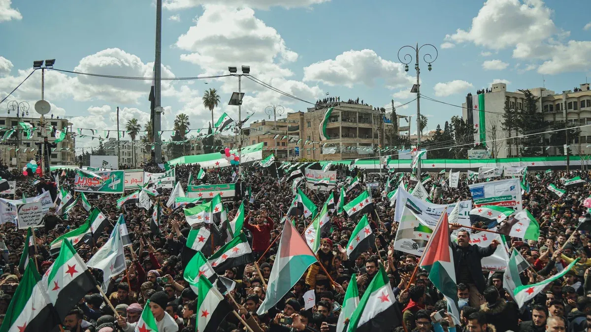 Crowd of people in a Syrian city square holding flags and banners during a peaceful demonstration.
