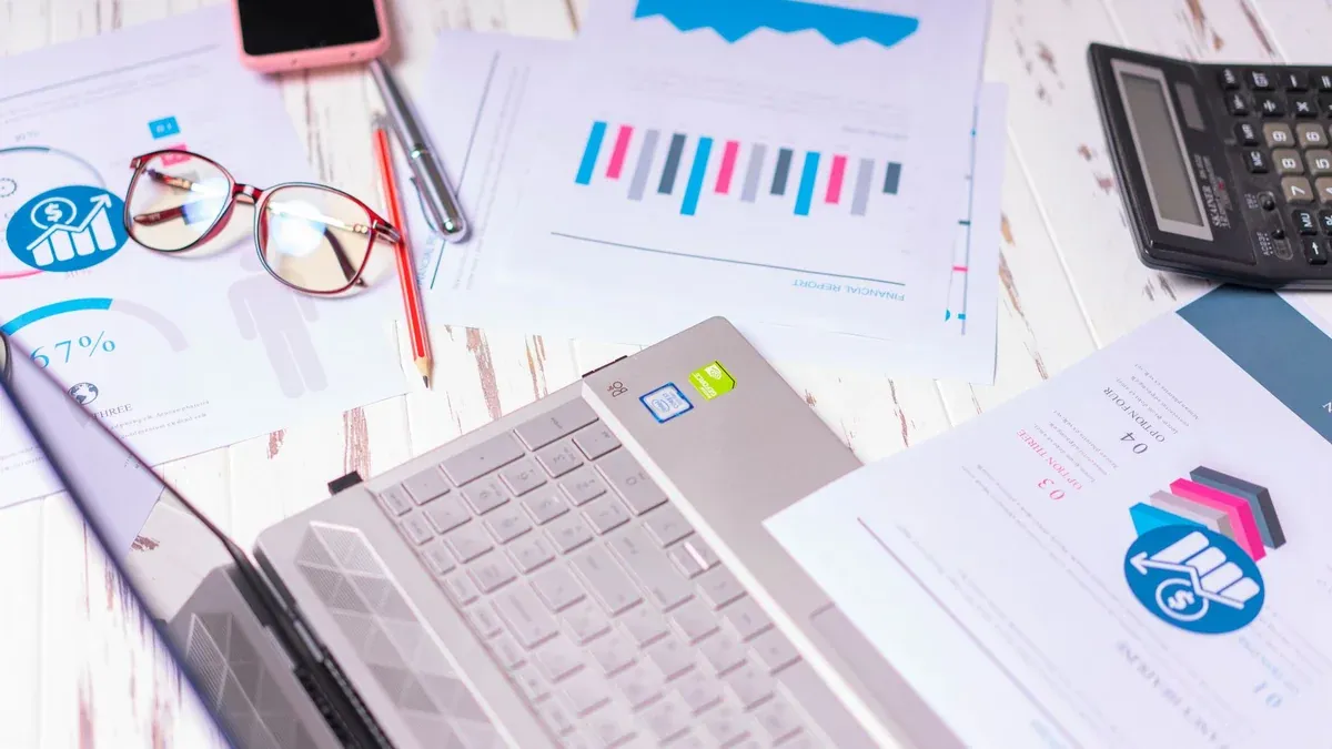 A cluttered desk with a laptop, financial reports, calculator, and glasses indicating a busy work environment.