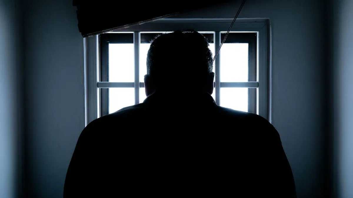 Dark silhouette of a man in a prison cell, backlit by window bars.