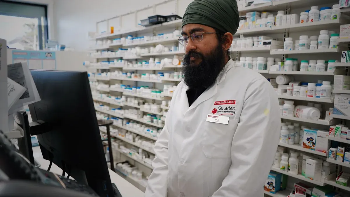 Pharmacist wearing a turban working at a computer in a pharmacy store with shelves filled with medicine.