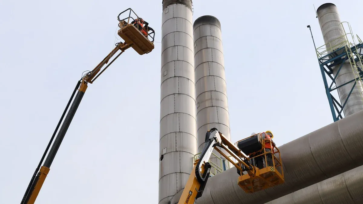 Workers using lifts for maintenance near towering factory chimneys against a clear sky.