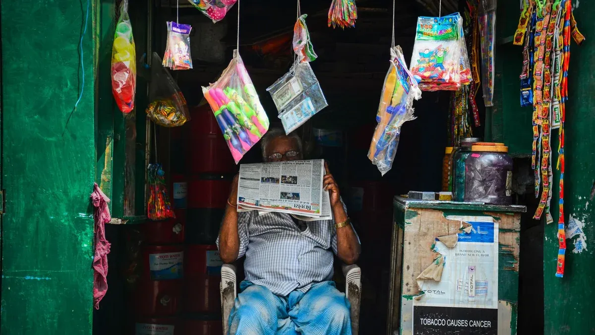 An elderly man reading a newspaper in a vibrant market shop filled with hanging goods.