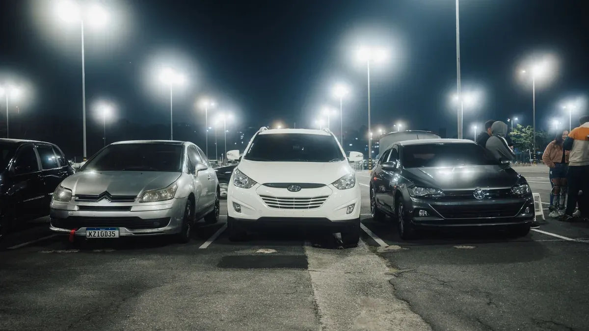 Night scene in a well-lit parking lot featuring various parked cars and street lamps.