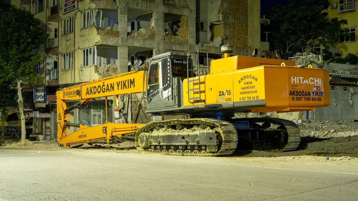 Large excavator at a city demolition site during nighttime, illuminated by streetlights.
