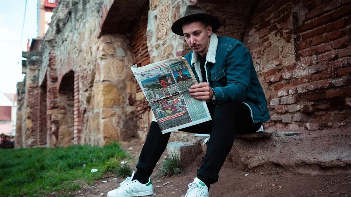 Young man in casual wear reading a newspaper against an ancient brick wall outdoors.