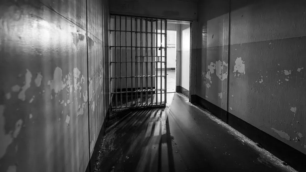 Moody black and white photo of a prison cell with an open barred door, evoking solitude.