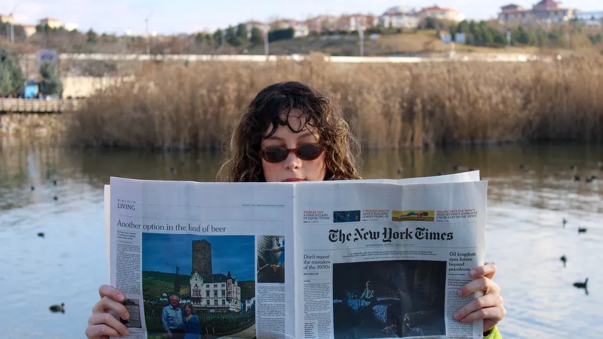 Woman with sunglasses reading a newspaper outdoors by a lake.