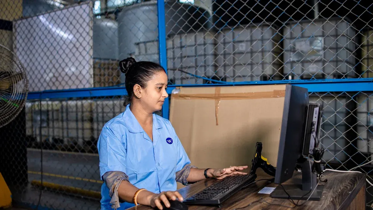 A woman in blue uniform operates a computer inside a factory warehouse, focusing on her task.