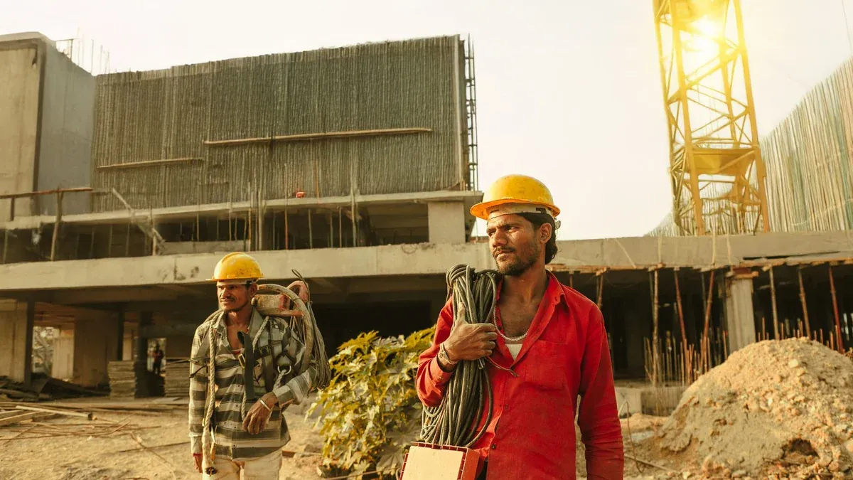 Two construction workers wearing helmets at a site in India with a crane and building.