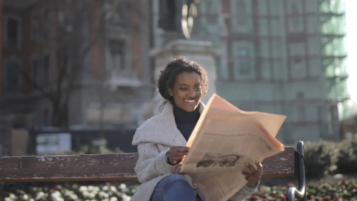 Cheerful woman enjoys reading a newspaper while seated on a park bench during a sunny day.