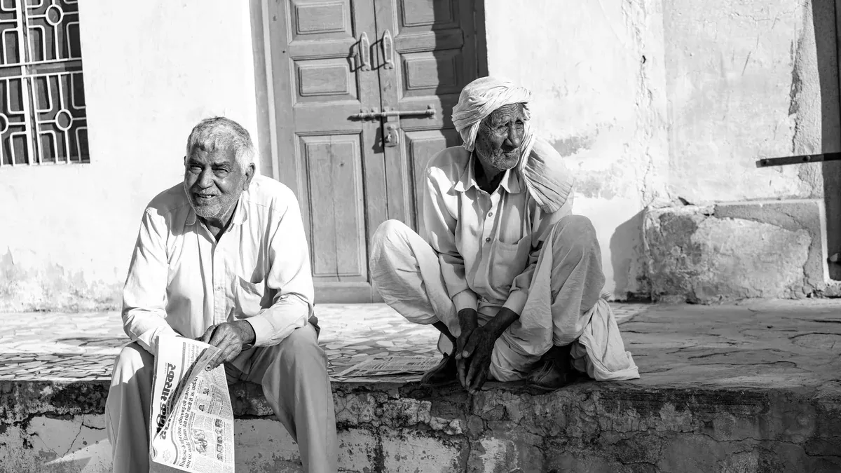 Two senior men relaxing outdoors, one reading a newspaper, captured in black and white for a nostalgic effect.