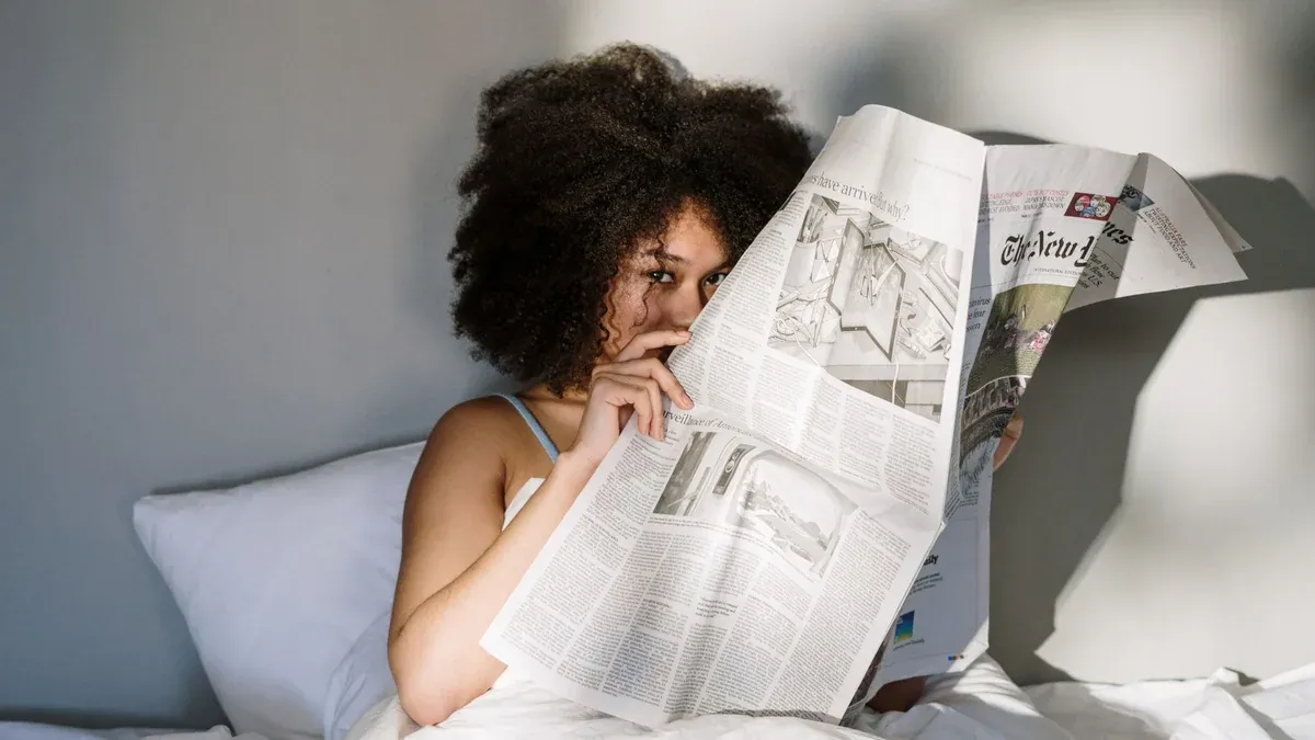 A relaxed morning scene of a woman reading a newspaper in bed, sunlight streaming in.
