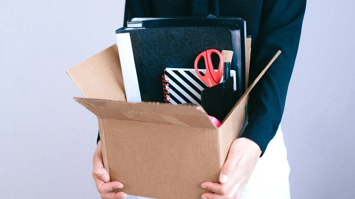 Woman carrying cardboard box with office items like scissors and notebooks.