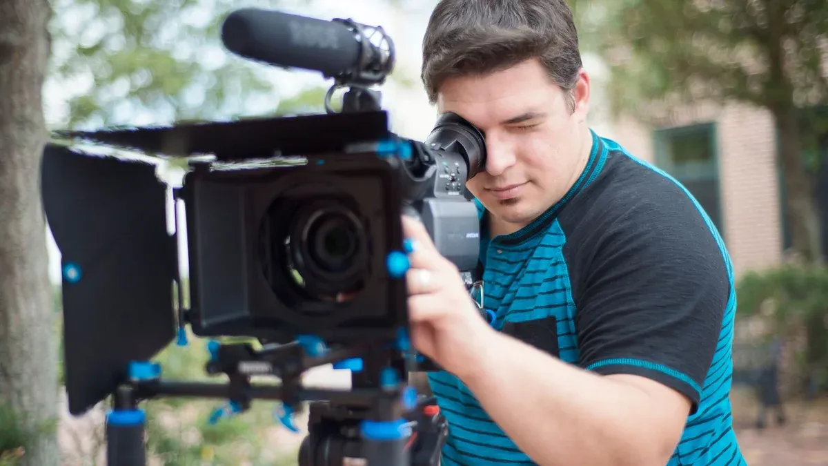 Young male videographer using a professional video camera outdoors with a blurred background.