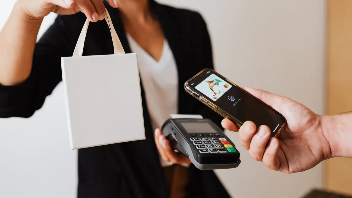 A person uses a smartphone for contactless payment with a payment terminal, while receiving a shopping bag.