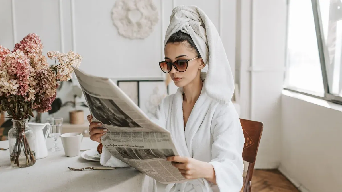 Woman in bathrobe reading a newspaper at a stylish indoor setting with flowers.