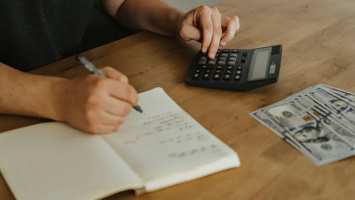 Close-up of hands using a calculator with notebook and cash on a wooden table.