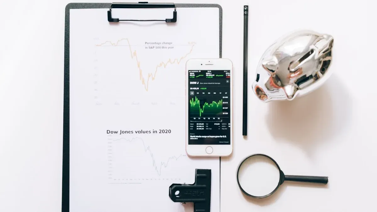 Top view of financial analysis tools including a cellphone, clipboard, magnifying glass, and piggy bank on a white desk.