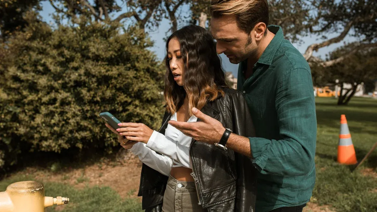 A couple having a disagreement outdoors, focusing on a cellphone. Emotions are visible.