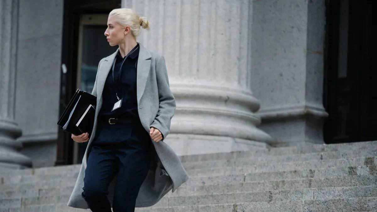 Serious female lawyer in elegant outfit holding folders with documents while going downstairs near stone state building and looking away