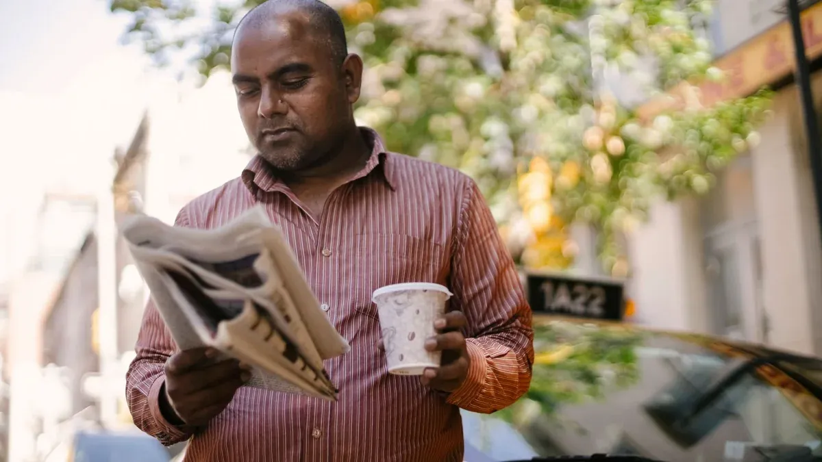 Interested adult ethnic male taxi driver with disposable glass of hot drink reading newspaper near vehicle on city street