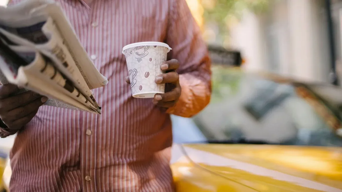 Crop anonymous ethnic male cab driver reading newspaper while standing with disposable glass of hot drink near polished vehicle on street
