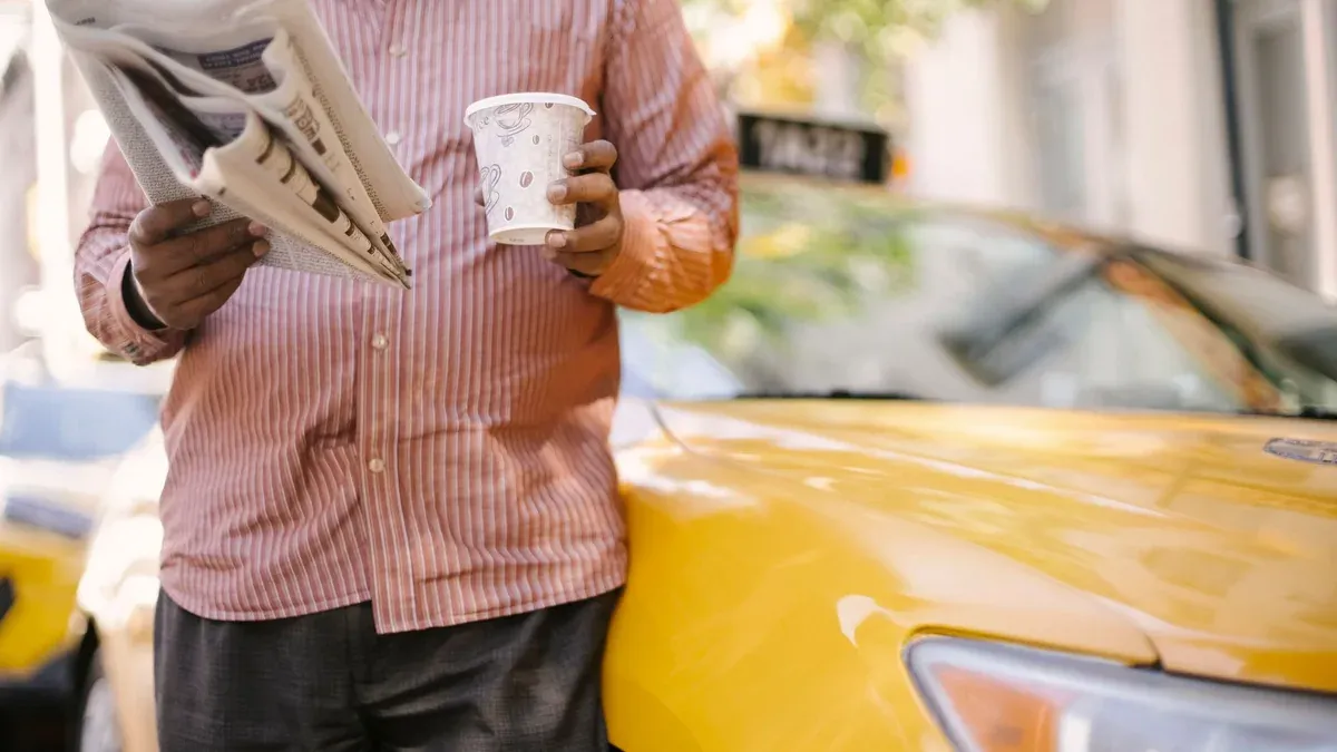 Crop unrecognizable ethnic male cab driver with plastic glass of beverage reading newspaper near contemporary car in town