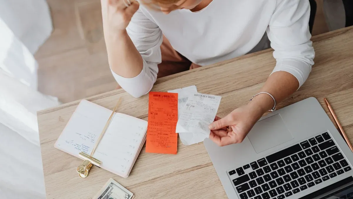 Woman reviewing receipts and planning budget using a laptop and notebook at home to manage expenses.