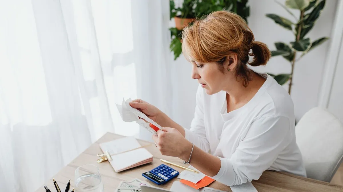 Woman sitting at a desk managing finances with a calculator and paper receipts in an office setting.