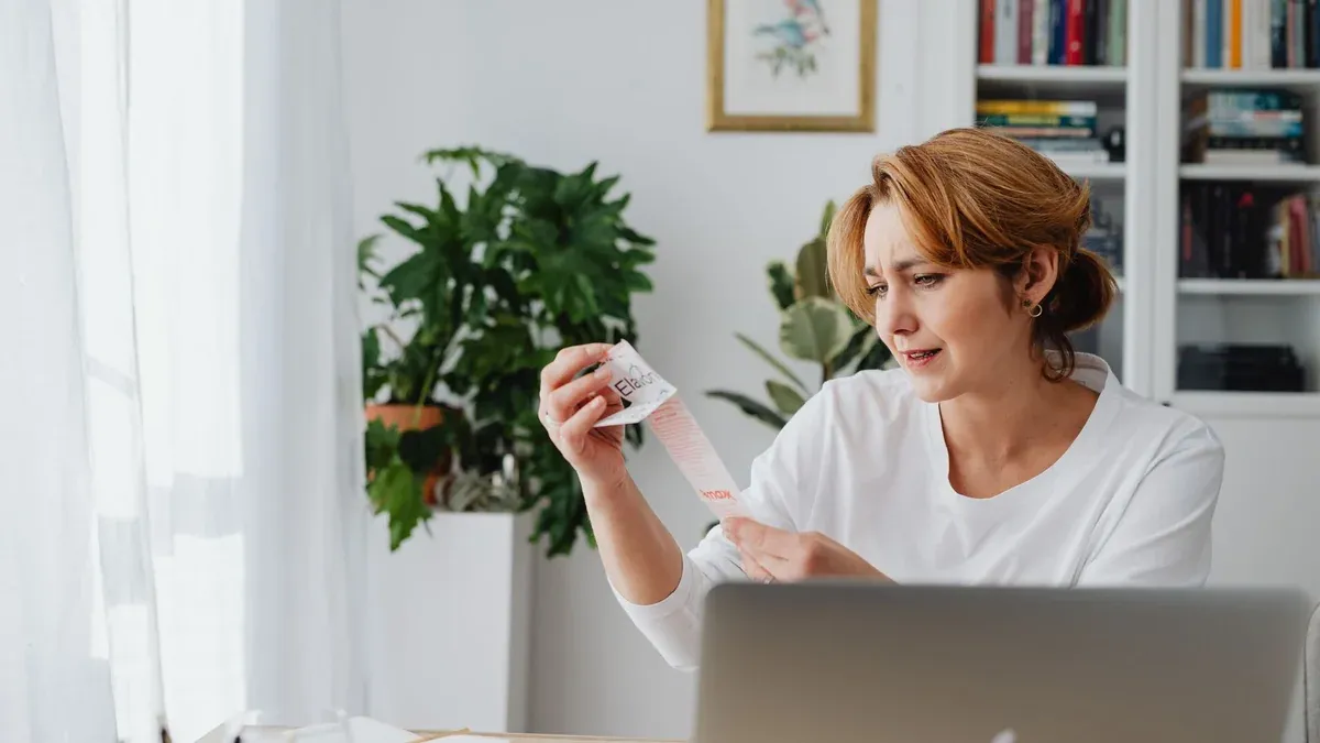 A woman in a white shirt sits indoors, examining a lengthy receipt with a concerned expression.