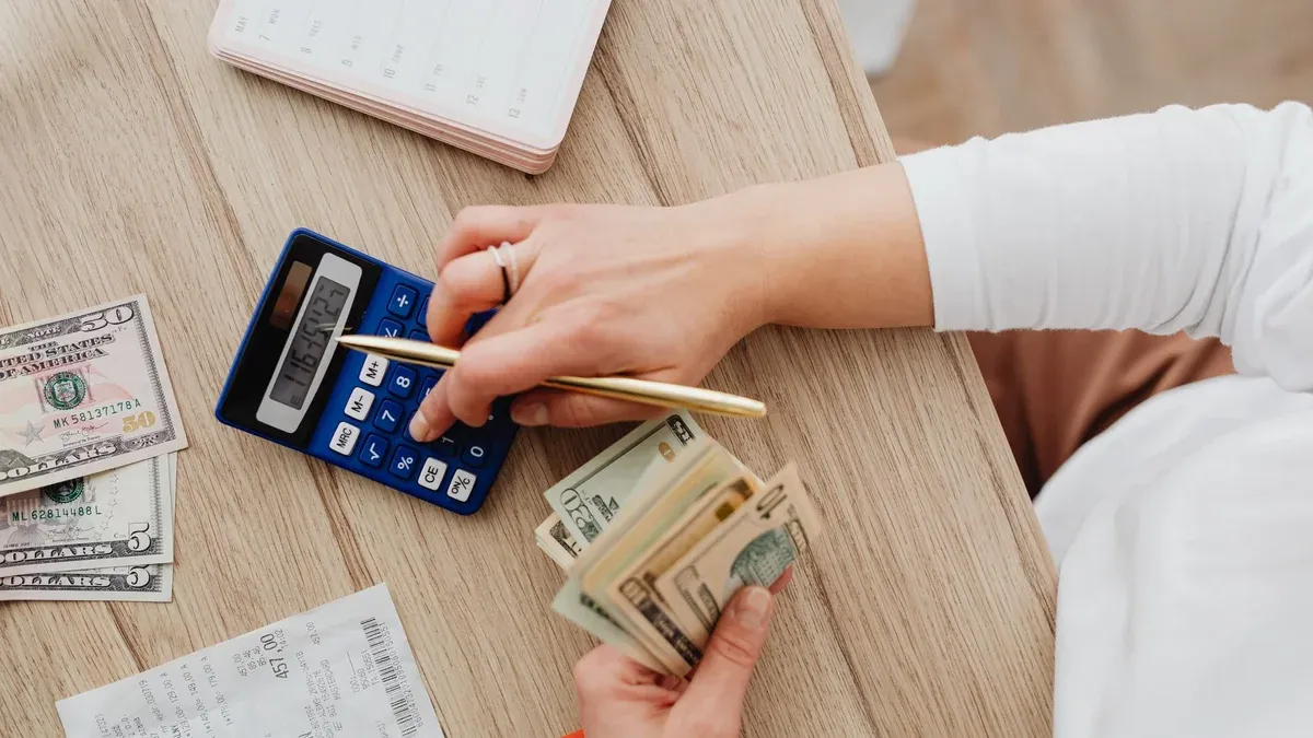 Person using a calculator and counting cash on a wooden table with receipts.