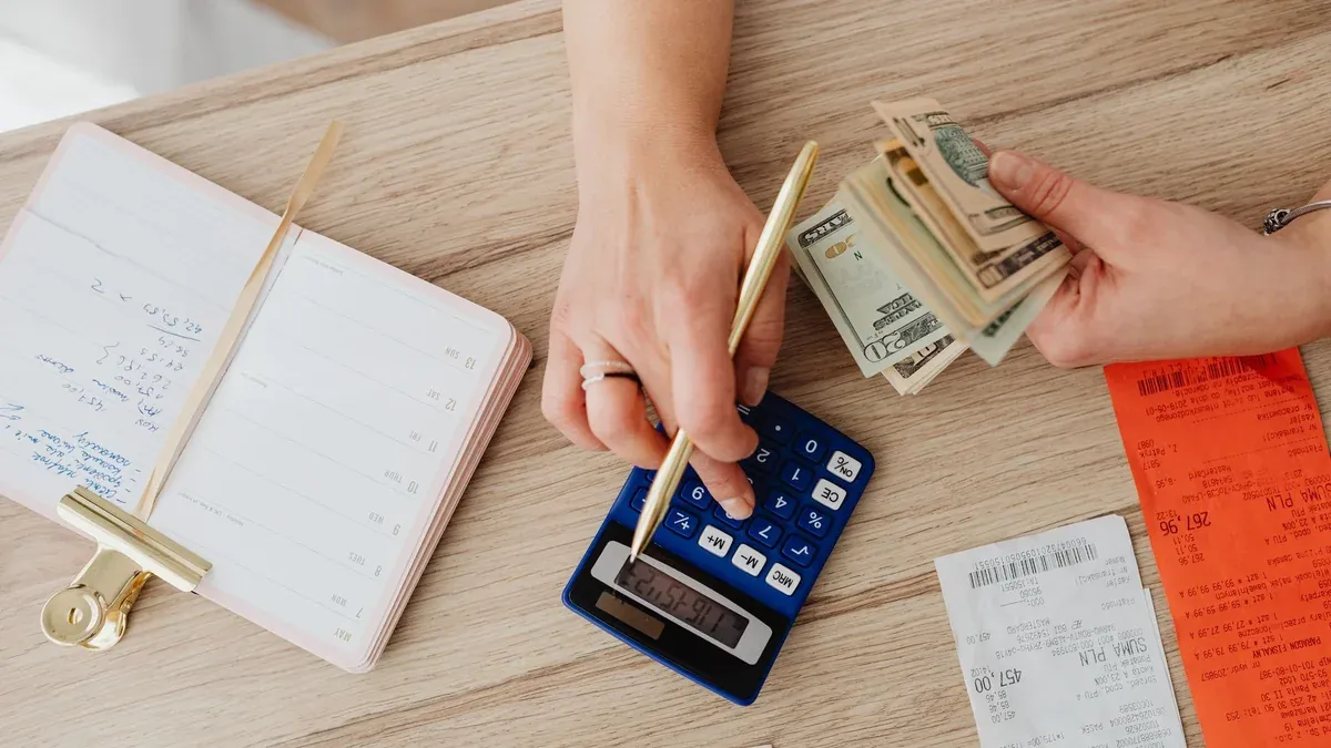 Hands managing finances with calculator, cash, and receipts on a wooden table. Ideal for budgeting concepts.
