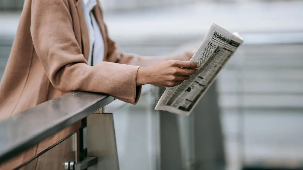 Side view of crop anonymous young African American female entrepreneur in stylish brown coat reading newspaper leaning on railing near blurred stairs