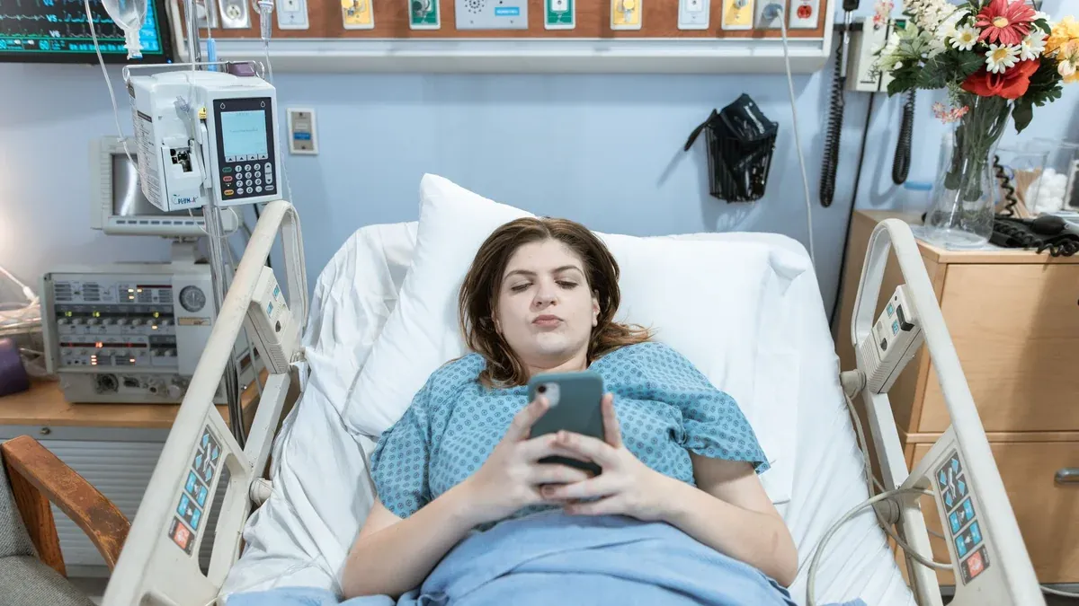 A female patient in a hospital bed using her phone, surrounded by medical equipment.