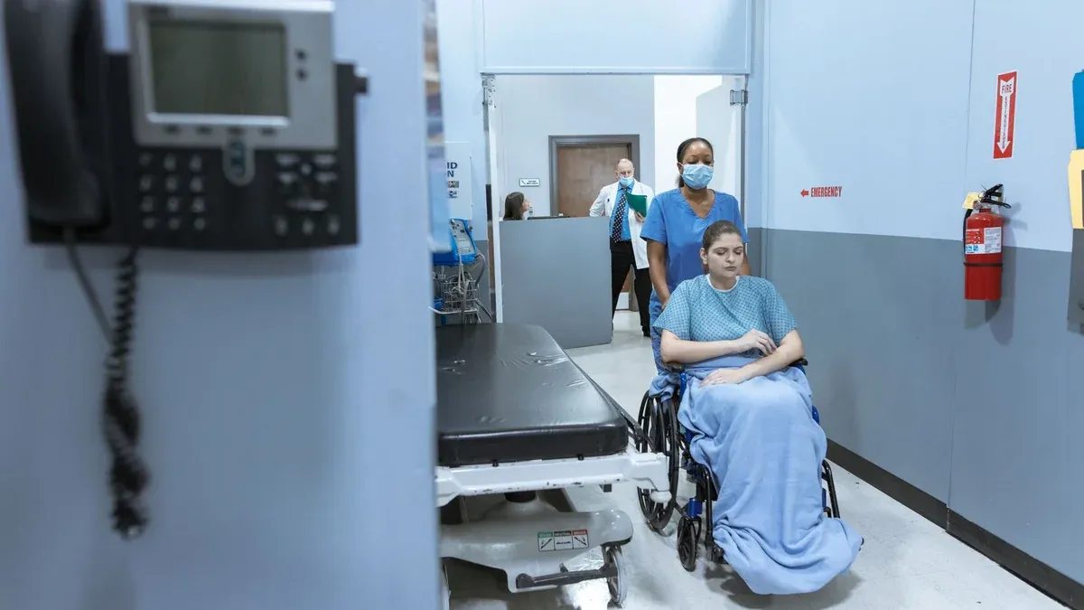 A nurse helps a patient in a wheelchair down a hospital corridor, reflecting care and medical professionalism.