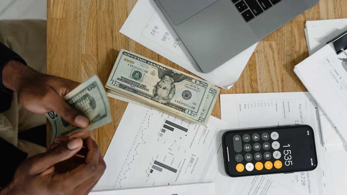 Close-up of hands counting cash on desk with calculator, charts, and laptop, illustrating financial management.