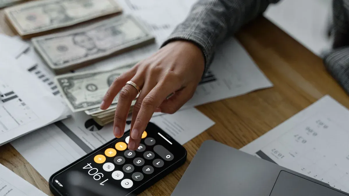 Close-up of a person using a smartphone calculator amid money and financial documents on a wooden table.