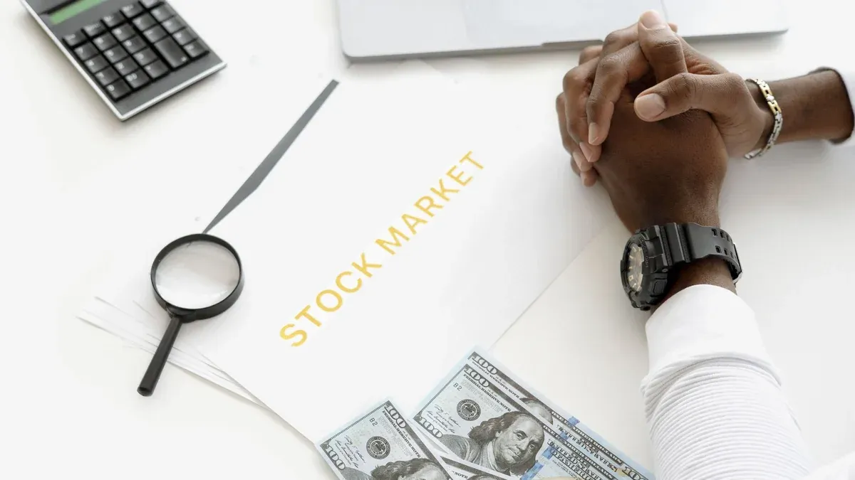 Hands folded over stock market documents with calculator and cash on desk.