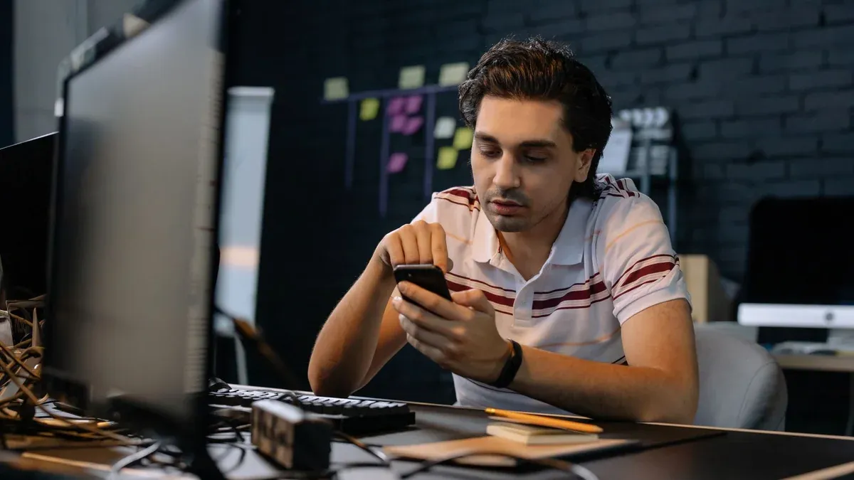 Young man in modern office using smartphone at his desk, focusing on multitasking work.