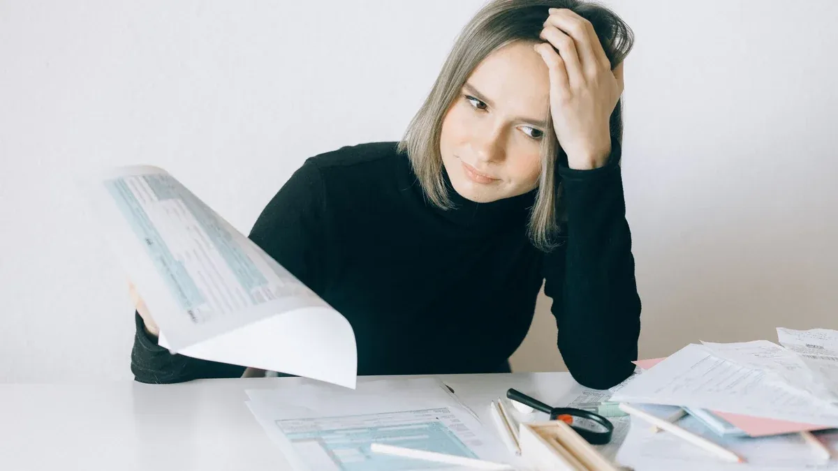 A woman showing stress while reviewing multiple paperwork and financial documents at a desk.