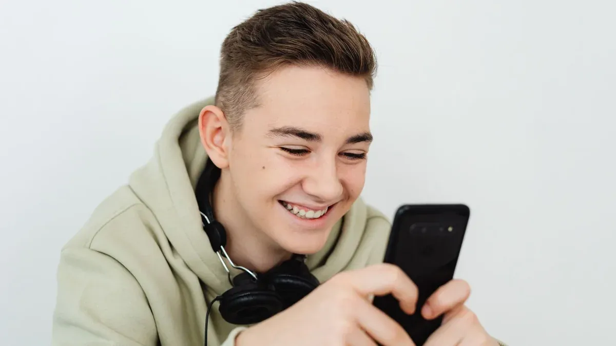 Smiling teenager using smartphone with headphones around neck indoors.