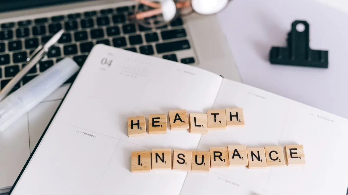 Close-up of Scrabble tiles spelling 'Health Insurance' on a planner with a laptop.