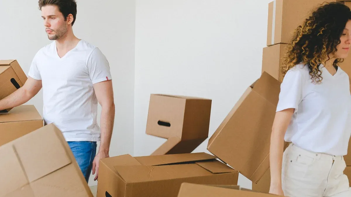 Serious young man and woman in white t shirts carrying carton boxes in light room while moving into new apartment together