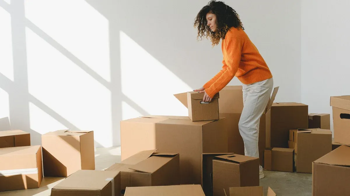 Side view of concentrated young lady with curly hair in casual clothes packing and arranging cardboard boxes while preparing for moving into new apartment in daylight